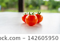 Fresh ripe red tomatoes on bright white tabletop with soft green garden blur, natural daylight, clean minimal food composition, healthy organic produce, close view, shallow focus, simple rustic 142605950