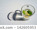 Clear glass of citrus water with lemon slice and mint leaves casting strong sunlit shadow on bright white surface, minimal summer refreshment composition with clean modern look 142605953