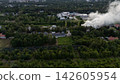 Aerial view of factory complex releasing heavy smoke over green industrial park, rural valley fields, and nearby homes beside warehouse roofs and parked vehicles 142605954