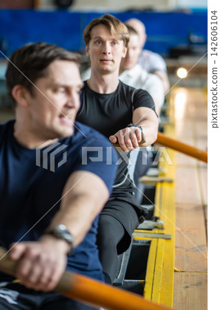 Focused male rowing crew pulling with strength during demanding indoor endurance training session Focused male rowing crew pulling with strength during demanding indoor endurance training session 142606104