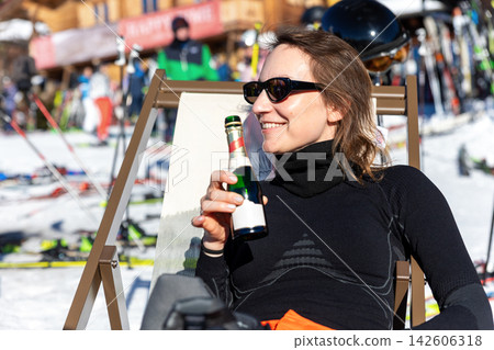 Smiling woman in sunglasses relaxes on deck chair at snowy ski resort, holding beer bottle beside slopes and equipment. Sunny winter scene suggests apres ski leisure, vacation, sport, and celebration Smiling woman in sunglasses relaxes on deck chair at snowy ski resort, holding beer bottle beside slopes and equipment. Sunny winter scene suggests apres ski leisure, vacation, sport, and celebration 142606318