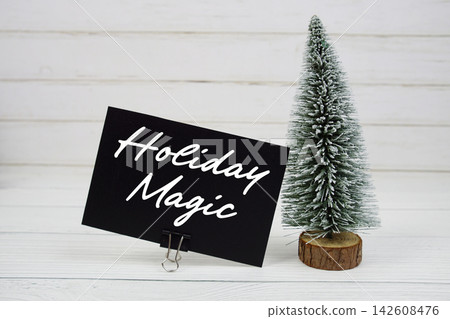 Holiday Magic text written on paper card with snowy tree on wooden background 142608476