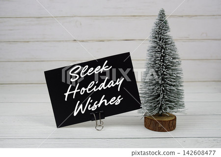 Sleek Holiday Wishes text written on paper card with snowy tree on wooden background Sleek Holiday Wishes text written on paper card with snowy tree on wooden background 142608477