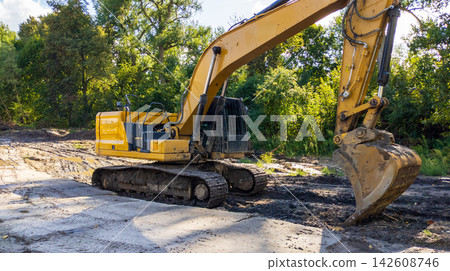 Yellow Excavator Digging Dirt on Construction Site with Big Bucket Machinery and Green Trees Background Yellow Excavator Digging Dirt on Construction Site with Big Bucket Machinery and Green Trees Background 142608746