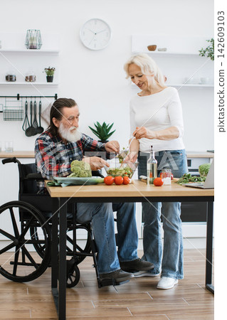 An elderly couple happily prepares a fresh salad together in a bright, modern kitchen, with the man in a wheelchair An elderly couple happily prepares a fresh salad together in a bright, modern kitchen, with the man in a wheelchair 142609103