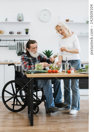 An elderly couple, one in a wheelchair, happily prepares a fresh salad in a bright, modern kitchen, showcasing teamwork and healthy living An elderly couple, one in a wheelchair, happily prepares a fresh salad in a bright, modern kitchen, showcasing teamwork and healthy living 142609104