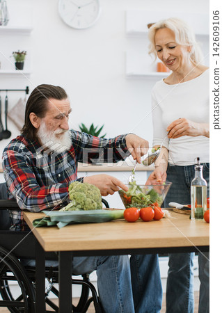 An elderly couple happily prepares a fresh salad together in a bright, modern kitchen, with the man seated in a wheelchair An elderly couple happily prepares a fresh salad together in a bright, modern kitchen, with the man seated in a wheelchair 142609106