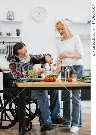 An elderly couple, one in a wheelchair, happily prepares a fresh salad together in a bright, modern kitchen, showcasing teamwork and healthy living An elderly couple, one in a wheelchair, happily prepares a fresh salad together in a bright, modern kitchen, showcasing teamwork and healthy living 142609107