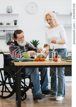 An elderly couple, one in a wheelchair, happily prepares a fresh salad in their bright, modern kitchen An elderly couple, one in a wheelchair, happily prepares a fresh salad in their bright, modern kitchen 142609108