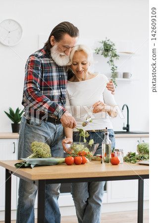 An elderly couple lovingly prepares a fresh, vibrant salad in their bright, modern kitchen, showcasing a healthy lifestyle 142609109
