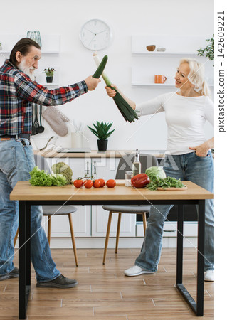 An older couple playfully pretends to fence with a cucumber and a leek in a bright, modern kitchen filled with fresh produce An older couple playfully pretends to fence with a cucumber and a leek in a bright, modern kitchen filled with fresh produce 142609221