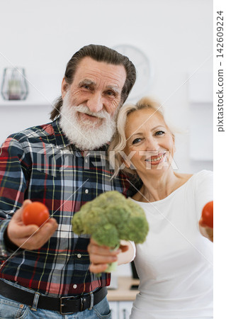 A joyful elderly couple smiles while holding fresh broccoli and tomatoes, embodying a healthy lifestyle and shared happiness in the kitchen A joyful elderly couple smiles while holding fresh broccoli and tomatoes, embodying a healthy lifestyle and shared happiness in the kitchen 142609224