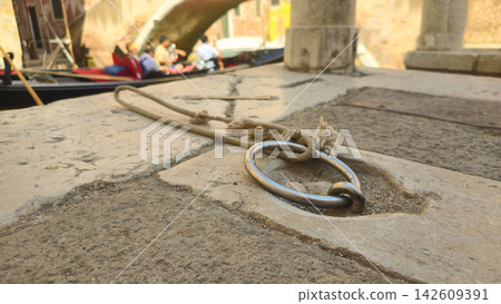 Metal mooring ring and rope sit beside a gondola on a canal in Venice, Italy. Concepts: Venetian travel, maritime heritage, transportation, tourism, traditional boating, urban waterways. Metal mooring ring and rope sit beside a gondola on a canal in Venice, Italy. Concepts: Venetian travel, maritime heritage, transportation, tourism, traditional boating, urban waterways. 142609391