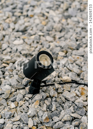 Black outdoor spotlight stands among gravel stones. Concept of landscape lighting, electricity, infrastructure, safety, visibility and outdoor design Black outdoor spotlight stands among gravel stones. Concept of landscape lighting, electricity, infrastructure, safety, visibility and outdoor design 142609733