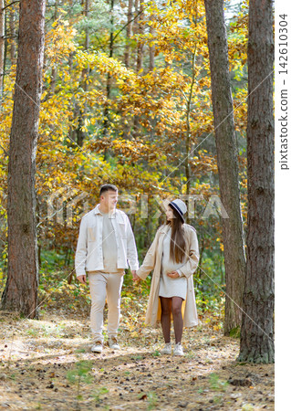 A pregnant woman and her partner stroll hand-in-hand through a beautiful forest during the fall season A pregnant woman and her partner stroll hand-in-hand through a beautiful forest during the fall season 142610304