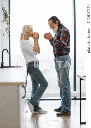 An elderly couple shares a warm moment over coffee cups in their bright, modern kitchen with a large window An elderly couple shares a warm moment over coffee cups in their bright, modern kitchen with a large window 142610384