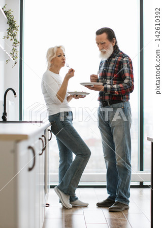 An older couple enjoys a meal together in their modern kitchen, standing by the counter near a large window An older couple enjoys a meal together in their modern kitchen, standing by the counter near a large window 142610392