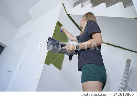 Woman paints interior wall green with roller while holding paint tray beside stairs Woman paints interior wall green with roller while holding paint tray beside stairs 142610860