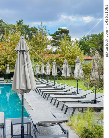 Resort Swimming Pool with Rows of Sun Loungers. Modern outdoor swimming pool with sun loungers, closed parasols, cloudy sky. Peaceful resort environment for relaxation, wellness, vacation travel Resort Swimming Pool with Rows of Sun Loungers. Modern outdoor swimming pool with sun loungers, closed parasols, cloudy sky. Peaceful resort environment for relaxation, wellness, vacation travel 142611063