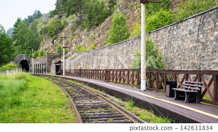 Rural railway station with a wooden platform and bench at Lake Baikal, Siberia. Train tracks curving towards a stone tunnel entrance in a green forested landscape. Railroad transportation concept Rural railway station with a wooden platform and bench at Lake Baikal, Siberia. Train tracks curving towards a stone tunnel entrance in a green forested landscape. Railroad transportation concept 142611834