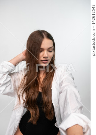 Young woman posing with long brown hair, wearing a white shirt and black bodysuit, showing natural beauty in a studio Young woman posing with long brown hair, wearing a white shirt and black bodysuit, showing natural beauty in a studio 142612542