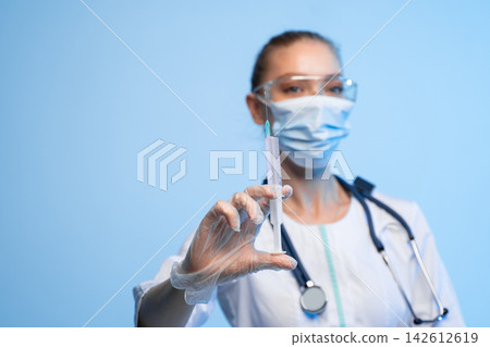Doctor or nurse wearing mask, safety glasses, and gloves, holding a syringe with vaccine or medication Doctor or nurse wearing mask, safety glasses, and gloves, holding a syringe with vaccine or medication 142612619