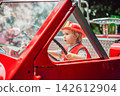Little boy dressed as firefighter playing in a red toy fire truck at an outdoor playground with helmet safety vest pretend rescue game childhood dreams role play and playing actively. 142612904