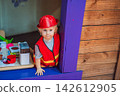 Little boy dressed as firefighter playing in a red toy fire truck at an outdoor playground with helmet safety vest pretend rescue game childhood dreams role play and rear view. 142612905
