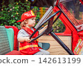 Little boy dressed as firefighter playing in a red toy fire truck at an outdoor playground with helmet safety vest pretend rescue game childhood dreams role play and walking. 142613199