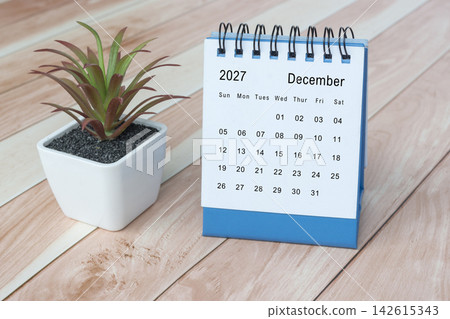 The December 2027 desk calendar with potted plant on wooden desk. The December 2027 desk calendar with potted plant on wooden desk. 142615343