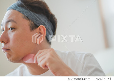 A 40-year-old man uses a gua sha plate to massage his neck and stimulate lymphatic drainage as part of his men's beauty routine. A 40-year-old man uses a gua sha plate to massage his neck and stimulate lymphatic drainage as part of his men's beauty routine. 142618031