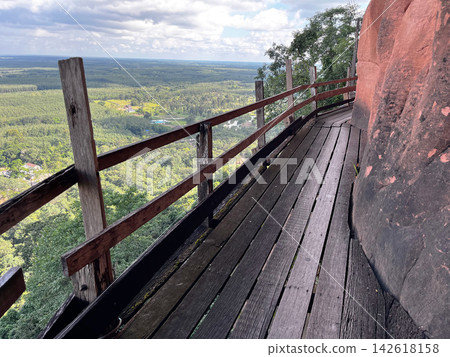 Wooden walkway around the steep sandstone cliff at Wat Phu Tok temple. Scenic mountain path trail with a wooden fence overlooking the green tropical forest and cloudy sky in Thailand Wooden walkway around the steep sandstone cliff at Wat Phu Tok temple. Scenic mountain path trail with a wooden fence overlooking the green tropical forest and cloudy sky in Thailand 142618158