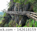 Wooden walkway around the steep sandstone cliff at Wat Phu Tok temple. Scenic mountain path trail with a wooden fence overlooking the green tropical forest and cloudy sky in Thailand 142618159