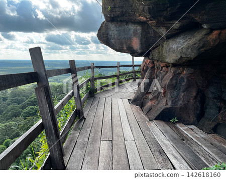 Wooden walkway around the steep sandstone cliff at Wat Phu Tok temple. Scenic mountain path trail with a wooden fence overlooking the green tropical forest and cloudy sky in Thailand Wooden walkway around the steep sandstone cliff at Wat Phu Tok temple. Scenic mountain path trail with a wooden fence overlooking the green tropical forest and cloudy sky in Thailand 142618160