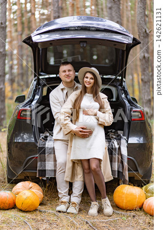 A happy couple poses in front of a car with pumpkins, celebrating their pregnancy in a beautiful outdoor setting A happy couple poses in front of a car with pumpkins, celebrating their pregnancy in a beautiful outdoor setting 142621131