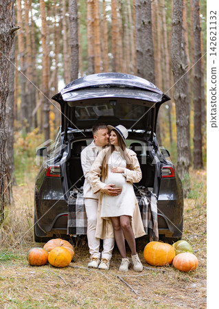 A loving couple expecting a baby embraces in a beautiful autumn forest, surrounded by pumpkins and a car A loving couple expecting a baby embraces in a beautiful autumn forest, surrounded by pumpkins and a car 142621132