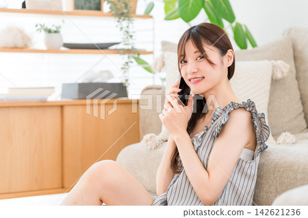 A young woman relaxes in a bright living room with her smartphone, enjoying her time at home with a smile. A young woman relaxes in a bright living room with her smartphone, enjoying her time at home with a smile. 142621236