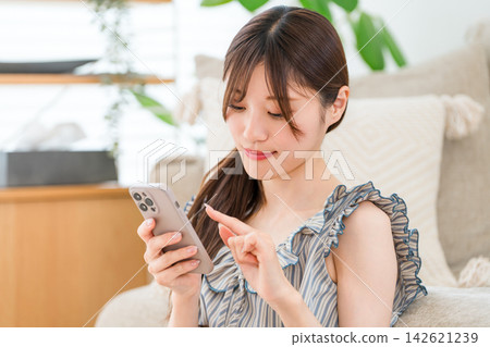 A young woman using her smartphone at home; enjoying her daily lifestyle of searching and shopping. A young woman using her smartphone at home; enjoying her daily lifestyle of searching and shopping. 142621239