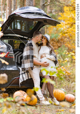A pregnant woman and her partner embrace near a car in a forest setting, surrounded by pumpkins and fall foliage A pregnant woman and her partner embrace near a car in a forest setting, surrounded by pumpkins and fall foliage 142623394