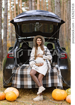 A beautiful pregnant woman poses in front of a car in a forest setting, surrounded by pumpkins and fall foliage A beautiful pregnant woman poses in front of a car in a forest setting, surrounded by pumpkins and fall foliage 142623416