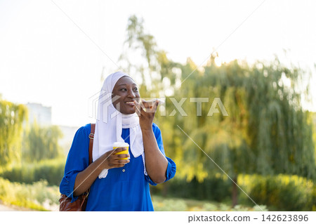 A smiling African American Muslim woman wearing a hijab holds a coffee cup while talking on her smartphone in a park setting A smiling African American Muslim woman wearing a hijab holds a coffee cup while talking on her smartphone in a park setting 142623896