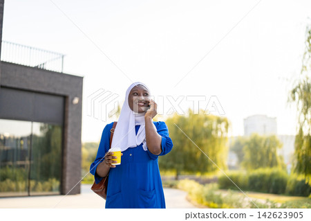A smiling African American Muslim woman in a hijab holds a coffee cup while talking on her phone outdoors A smiling African American Muslim woman in a hijab holds a coffee cup while talking on her phone outdoors 142623905