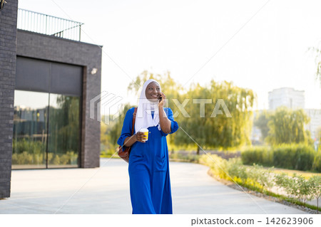 A smiling African American Muslim woman in a blue dress and white hijab walks outdoors while talking on her phone and holding a coffee cup 142623906