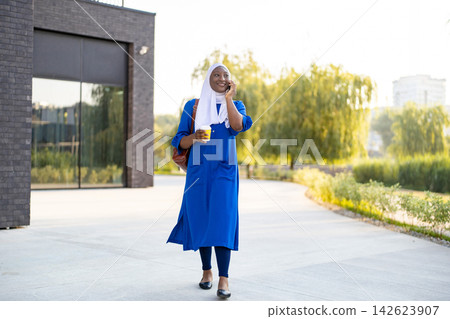 A smiling African American Muslim woman in a blue dress and hijab walks outdoors while talking on her phone and holding a coffee cup A smiling African American Muslim woman in a blue dress and hijab walks outdoors while talking on her phone and holding a coffee cup 142623907