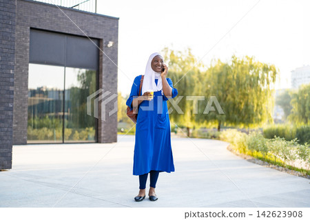 A smiling African American Muslim woman wearing a hijab and a blue dress walks while holding a coffee cup and talking on her phone A smiling African American Muslim woman wearing a hijab and a blue dress walks while holding a coffee cup and talking on her phone 142623908