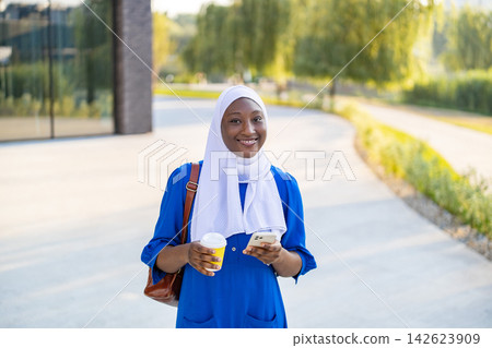 A happy African American Muslim woman in a white hijab and blue dress holds a coffee cup and smartphone while walking outdoors A happy African American Muslim woman in a white hijab and blue dress holds a coffee cup and smartphone while walking outdoors 142623909