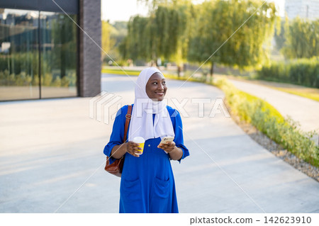 A smiling African American Muslim woman wearing a blue dress and white hijab walks outdoors with a coffee cup and smartphone in her hands A smiling African American Muslim woman wearing a blue dress and white hijab walks outdoors with a coffee cup and smartphone in her hands 142623910