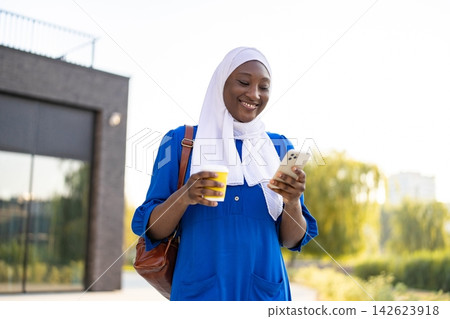 A happy African American Muslim woman in a blue dress and white hijab holds a coffee cup and a smartphone while walking outdoors A happy African American Muslim woman in a blue dress and white hijab holds a coffee cup and a smartphone while walking outdoors 142623918