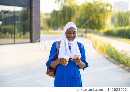 A smiling African American Muslim woman in a blue dress and white hijab walks outdoors holding a coffee cup and her smartphone A smiling African American Muslim woman in a blue dress and white hijab walks outdoors holding a coffee cup and her smartphone 142623922