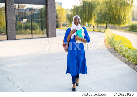 A happy African American Muslim woman wearing a hijab and a blue dress walks with books and a backpack outside a modern building A happy African American Muslim woman wearing a hijab and a blue dress walks with books and a backpack outside a modern building 142623929
